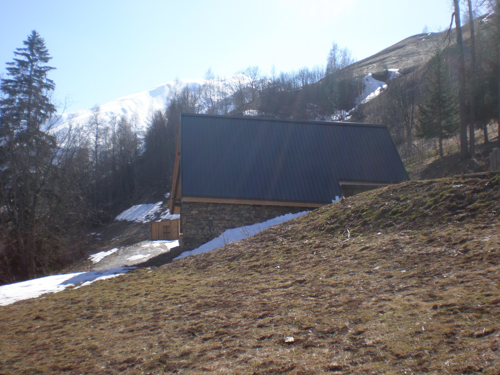 Stone building with a dark roof on a still snow-covered mountainside, peaks in the background
