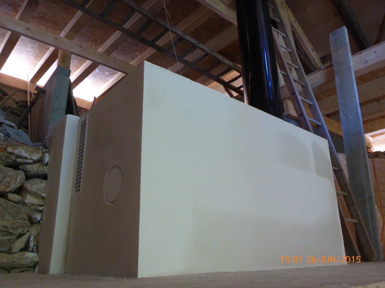 Upper part of the masonry heater emerging from the upstairs floor, rendered white, with a diffusion grille and a round cleaning door, beneath the roof frame
