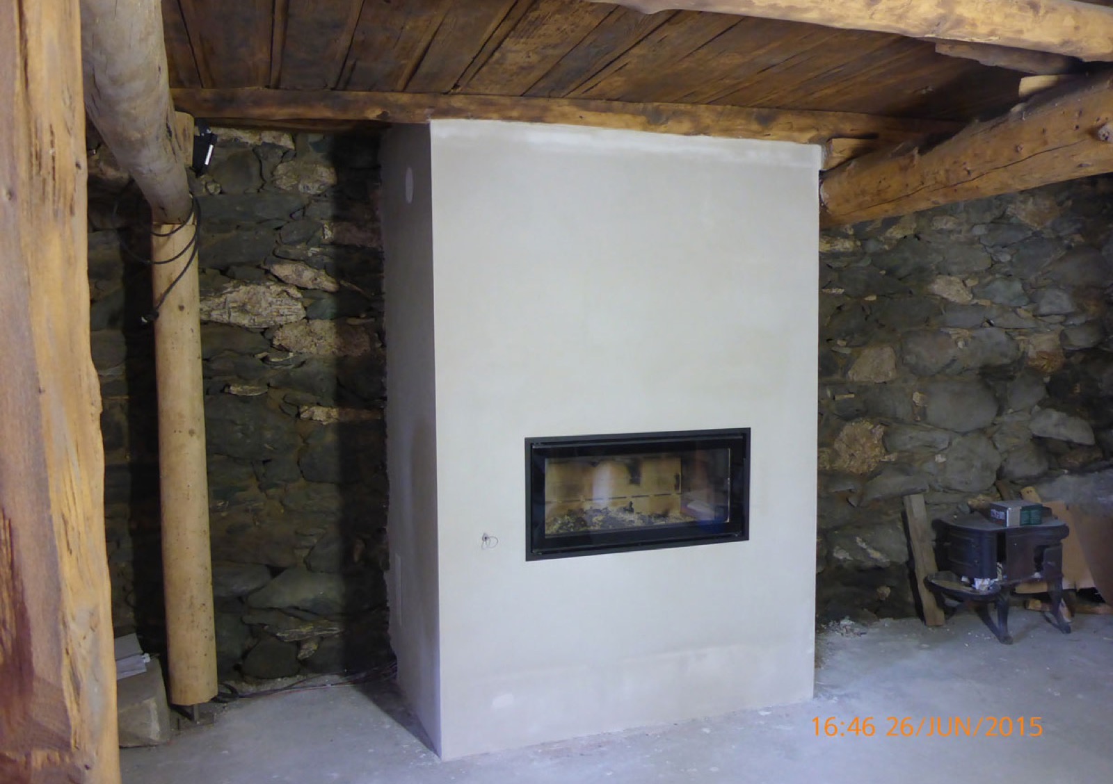 Cream-rendered masonry heater with a horizontal firebox, set against a dry-stone wall beneath a log roof frame