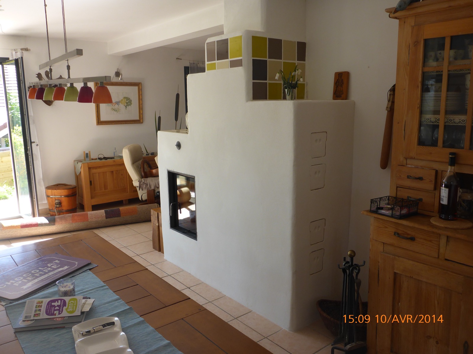 White rendered masonry heater with see-through firebox, crowned with a band of chequered tiles, between a kitchen and a living room