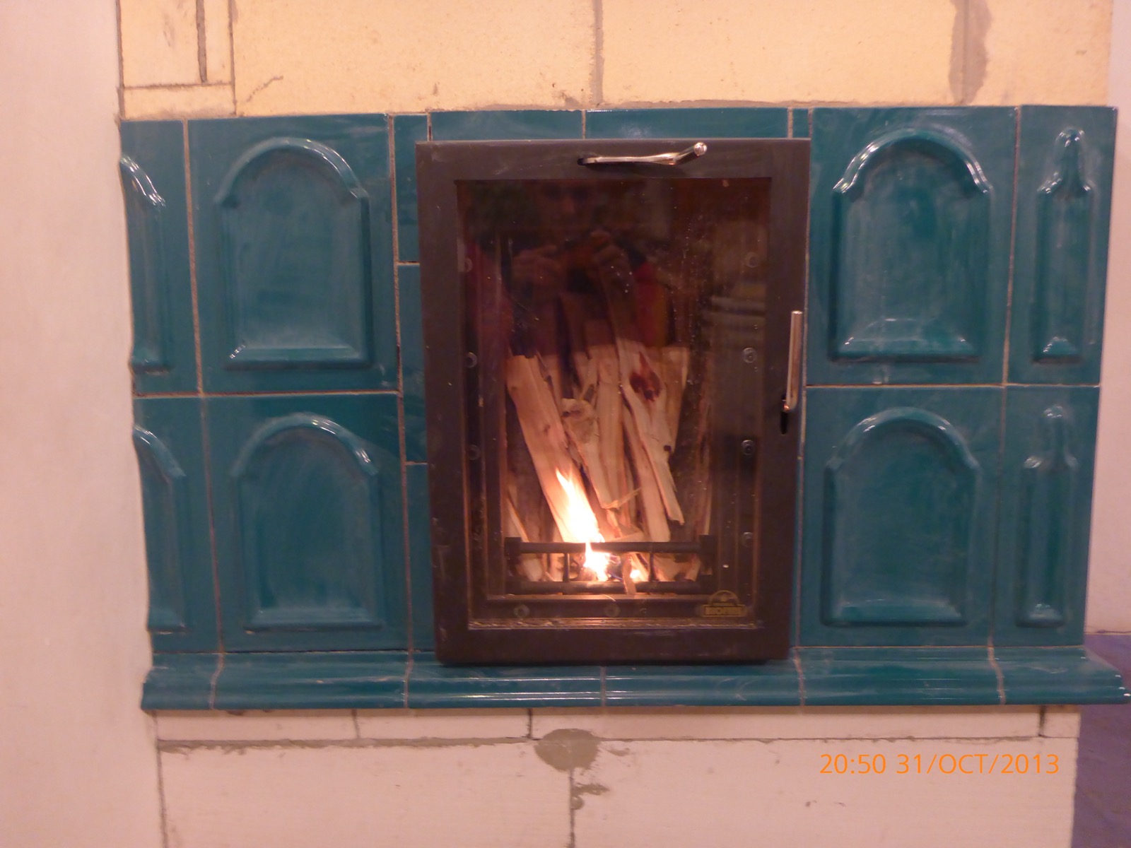 Primer plano del hogar encendido, enmarcado por paneles de azulejo verde azulado moldeados en arquería, puerta de bronce con tirador vertical