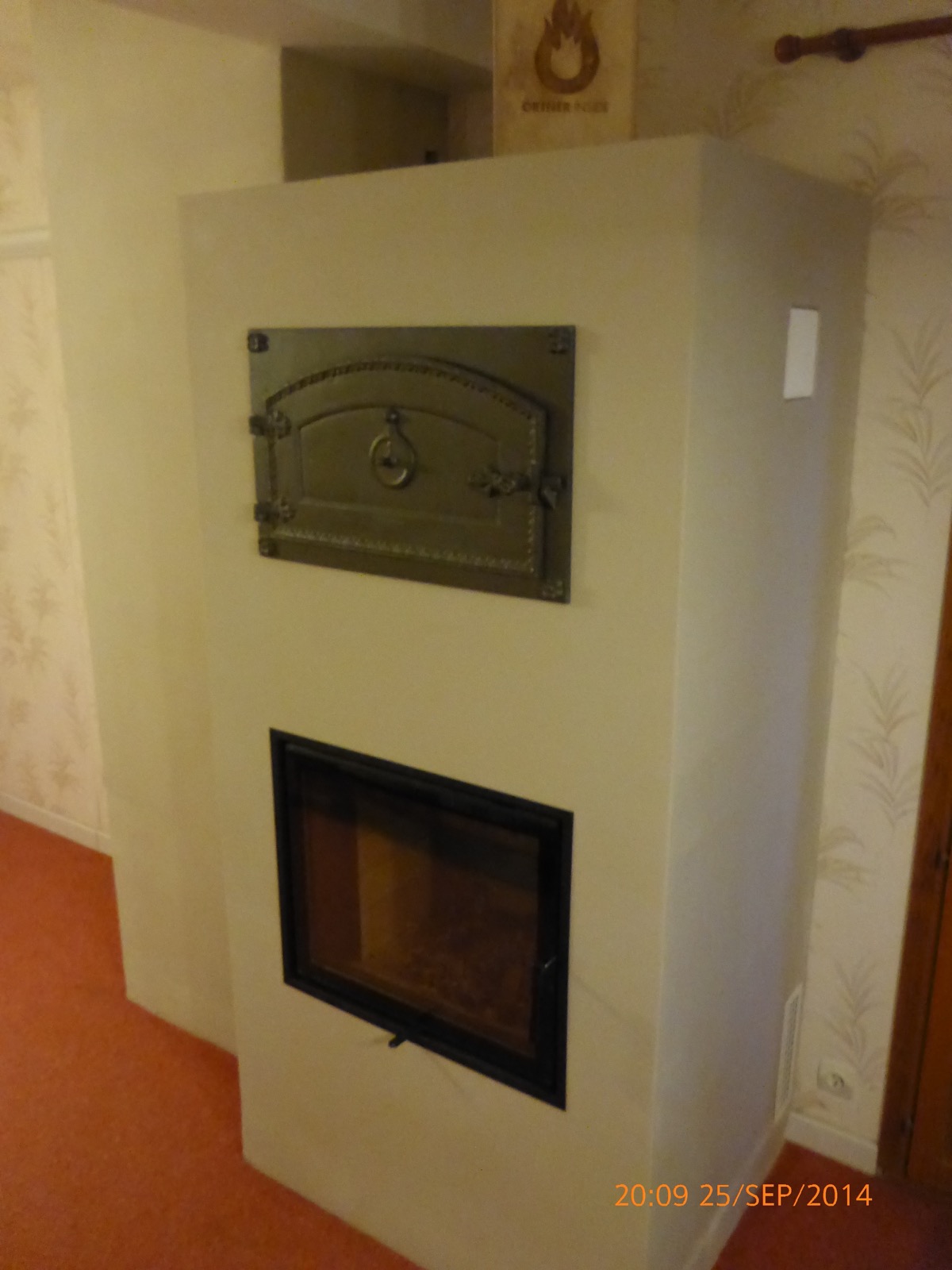 Cream-rendered masonry heater seen from three-quarters: glazed firebox at the bottom and ornate cast-iron oven door above