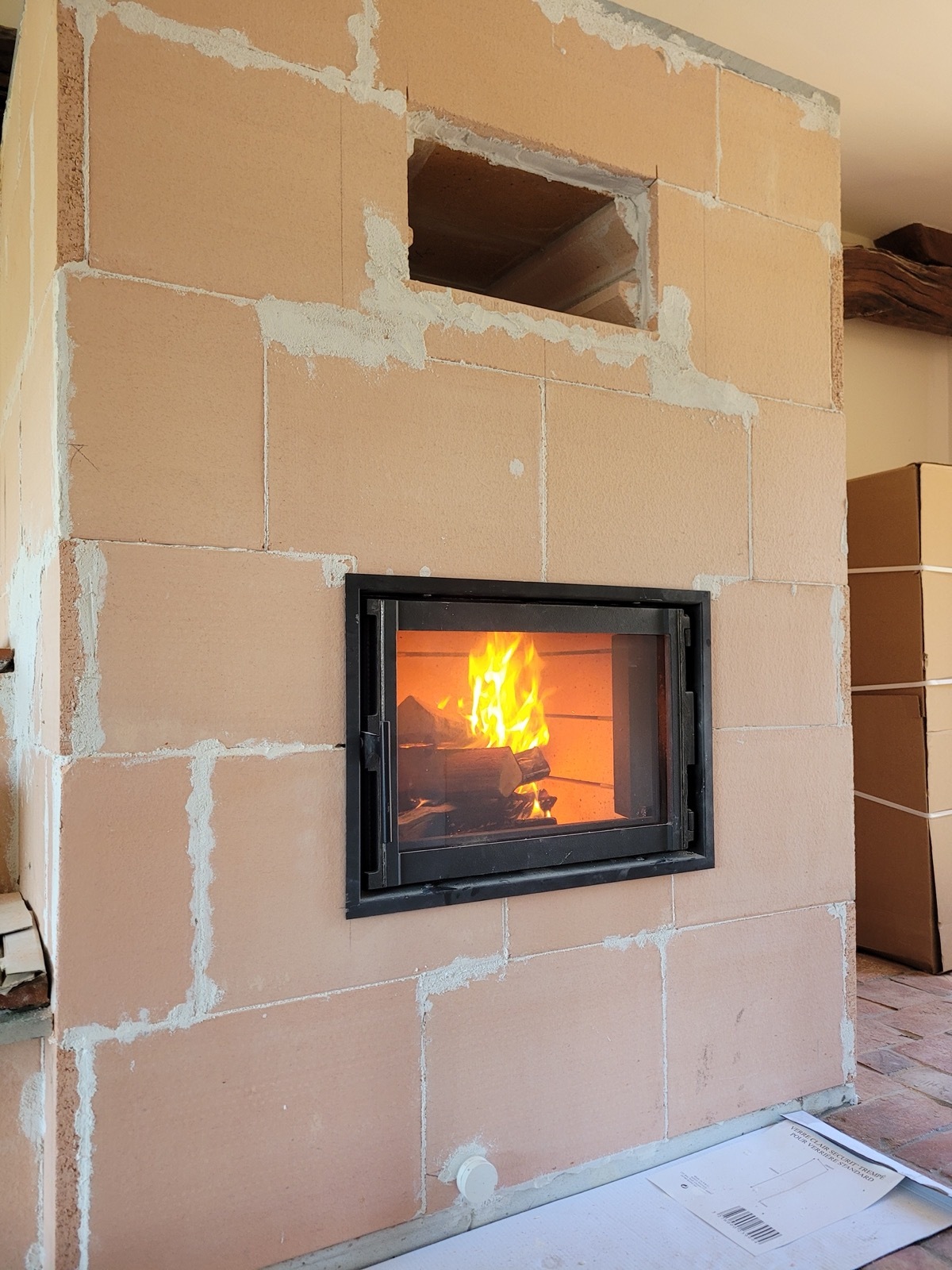Masonry heater built from pink refractory blocks with a see-through niche at the top, first fire lit in the glazed firebox