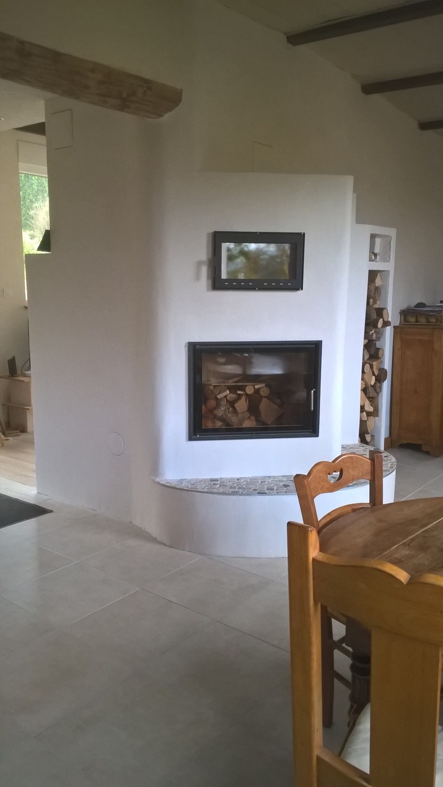 White-rendered Grundofen masonry heater with a rounded corner, glazed firebox, glazed oven door above, heated bench in pebbles and log niche