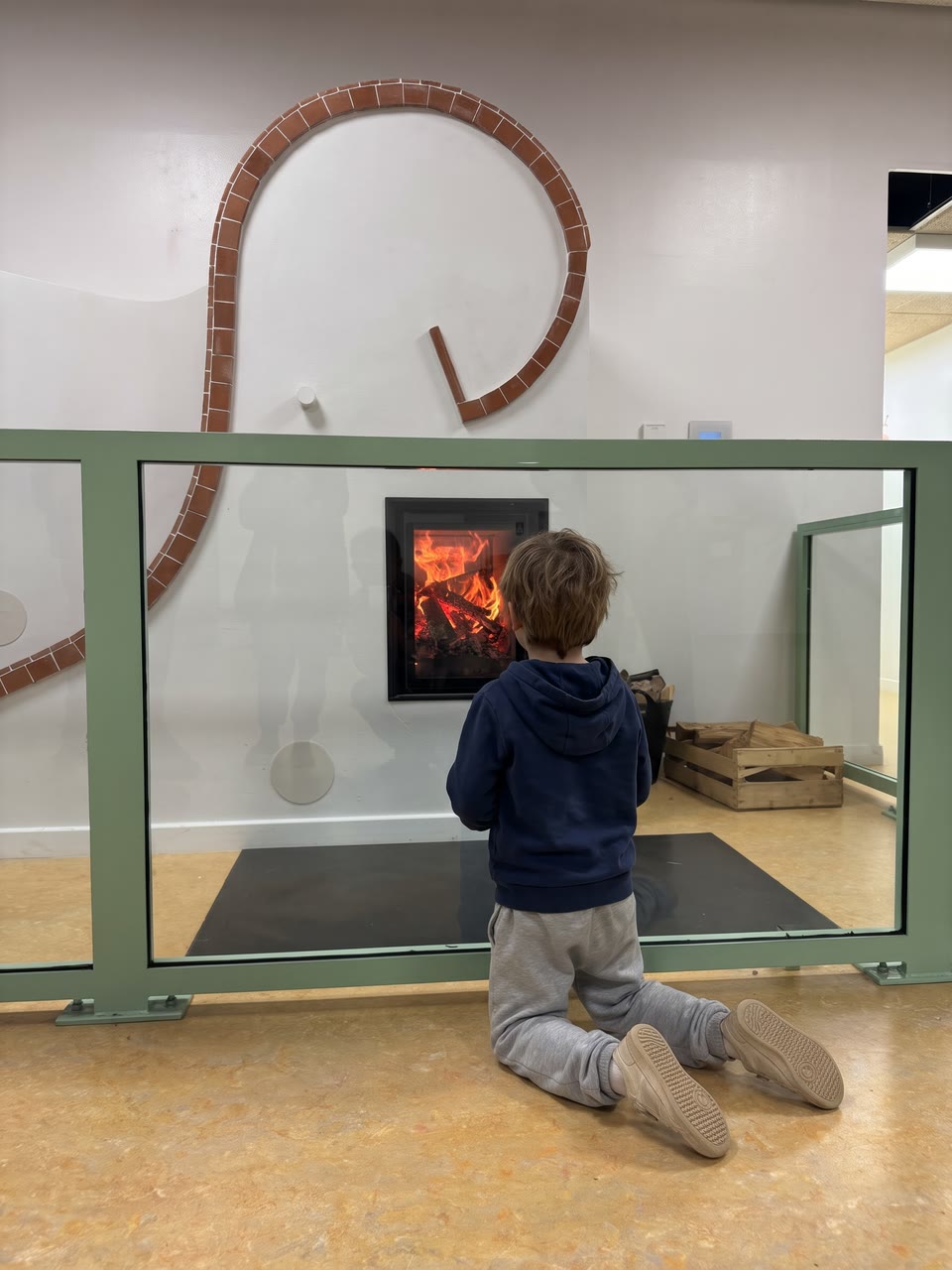 A child, seen from behind, watches the fire through the glass barrier of the Bois-Perrier school masonry heater