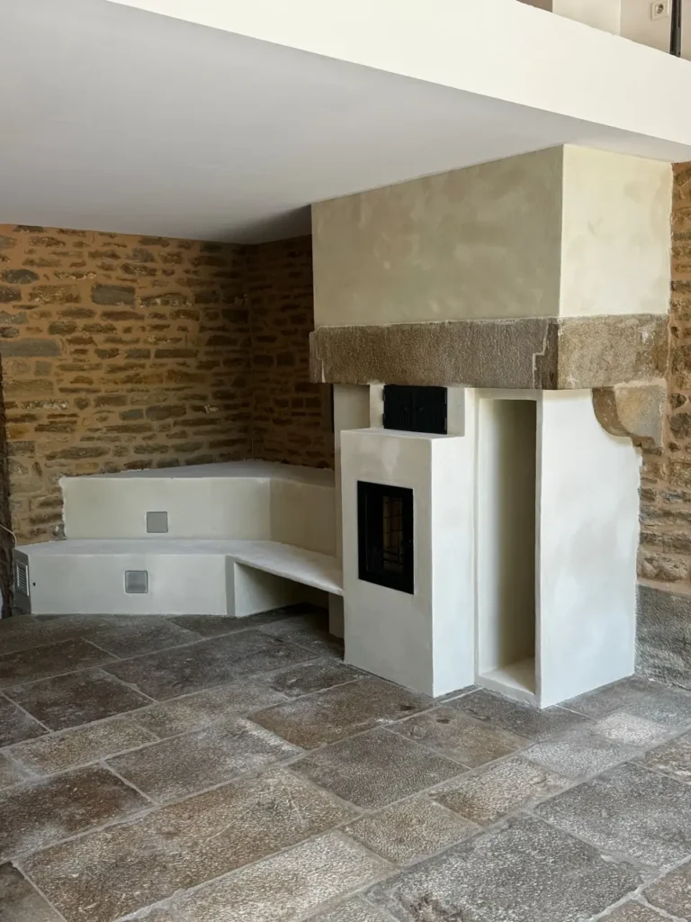 Masonry heater in the old fireplace: pale render, glazed firebox, log niche and corner bench, before the stone seats are fitted